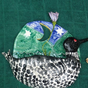 May include: A ceramic wall hanging depicting a loon with a black and white patterned body, a black head with a red eye, and a long neck. The loon is swimming in a blue and green wave with white stars and a crescent moon. A purple flower with a green stem is growing out of the wave.
