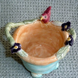 May include: A handmade ceramic bowl with a light orange interior and a turquoise base. The rim is decorated with green vines, purple flowers, and a red bird. The bowl has a handle and is likely used for decorative purposes.