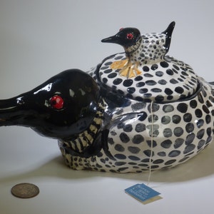 May include: A ceramic teapot shaped like a loon bird, featuring a black head and tail, red eyes, and a white body with black spots. A smaller bird sits on top. Includes a tea bag.