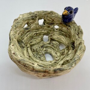 May include: A ceramic bird's nest bowl with a blue bird perched on the edge. The nest is textured with holes and a light green and brown glaze. The bird has a yellow beak.