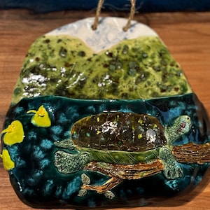 May include: A ceramic wall hanging featuring a turtle resting on a branch in a teal blue water scene. The top portion depicts green hills and a white and blue sky. A jute loop is attached for hanging.