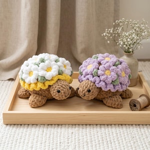 May include: Two handmade crochet turtle plushies. One has a white daisy shell with yellow and green accents, the other has a purple flower shell. Both have brown bodies and are on a wooden tray.