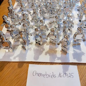 Op de afbeelding: Een verzameling glanzende, verchroomde speelfiguren, die lijken op miniatuurrobots of mensen, is gerangschikt op een wit oppervlak. De figuren zijn zeer reflecterend, met zichtbare details. Een handgeschreven notitie luidt "Chromebricks 16.09.25".