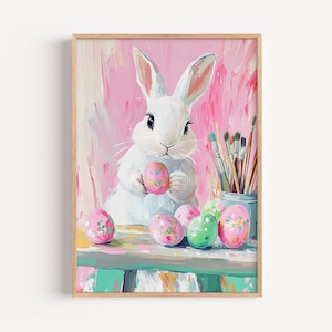 May include: A framed painting of a white rabbit holding two decorated Easter eggs. The rabbit is in front of a pink background. Several colorful Easter eggs and paintbrushes are on a table in front of the rabbit. The painting has a whimsical, artistic style.