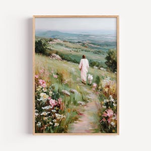 Op de afbeelding: Een ingelijste kunstdruk toont een persoon in een wit gewaad die over een pad door een veld met wilde bloemen loopt, gevolgd door een lam. De achtergrond toont glooiende groene heuvels en een verre bergketen onder een zachte, bewolkte lucht. Pastorale thema.