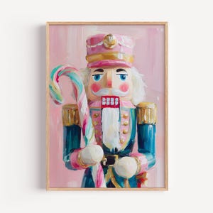 May include: A framed painting of a colorful nutcracker soldier holding a candy cane. The nutcracker wears a pink hat and jacket with gold accents. The background is a soft pink.