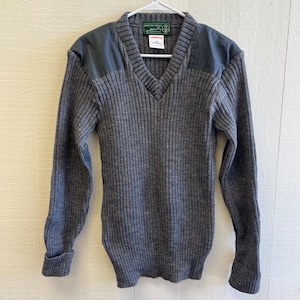 May include: A charcoal gray V-neck sweater featuring a ribbed knit texture, long sleeves, and solid dark gray fabric patches on the shoulders. The interior collar displays a green British Wool label and a white tag with text including 100% Pure New Wool.