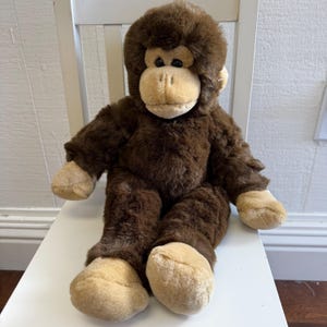 May include: A plush stuffed animal in the shape of a gorilla with dark brown fur and a light tan face, hands, and feet. The toy features round black eyes and a soft, rounded snout, with a seated posture and thick, textured synthetic fur.