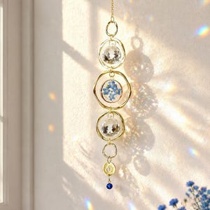 May include: A hanging suncatcher with a gold chain and circular frames. The suncatcher features clear crystal orbs and a central frame with blue flowers. A small blue bead hangs at the bottom. The suncatcher is hanging near a window.