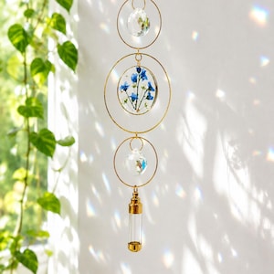 Könnte beinhalten: Ein hängender Sonnenfänger mit goldener Kette und kreisförmigen Rahmen. Der Sonnenfänger hat einen klaren Kristallkugel, einen Kreis mit blauen Blumen und ein kleines Glasröhrchen. Der Sonnenfänger hängt vor einer weißen Wand.