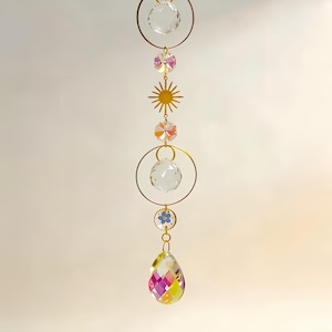 Könnte beinhalten: Ein dekorativer Sonnenfänger mit goldener Kette und Akzenten. Er verfügt über klare Kristallkugeln, einen Sonnenstrahl und einen tropfenförmigen Kristall mit rosa, gelben und violetten Facetten. Das Design beinhaltet eine kleine blaue Blume.