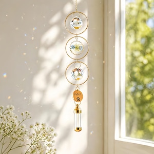 Könnte beinhalten: Ein hängendes Gedenk-Windspiel mit drei goldfarbenen Ringen. Zwei klare Kristallkugeln und ein Blumendesign sind in den Ringen aufgehängt. Ein Holzanhänger mit dem Namen "Peter" und einem Pfotenabdruck hängt darunter, zusammen mit einem Glasfläschchen.
