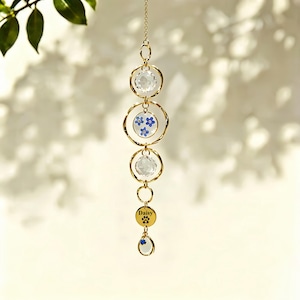 May include: A gold-coloured hanging suncatcher with clear crystal elements and blue floral patterns. The design includes a gold chain and a charm with the name "Daisy" and a paw print.