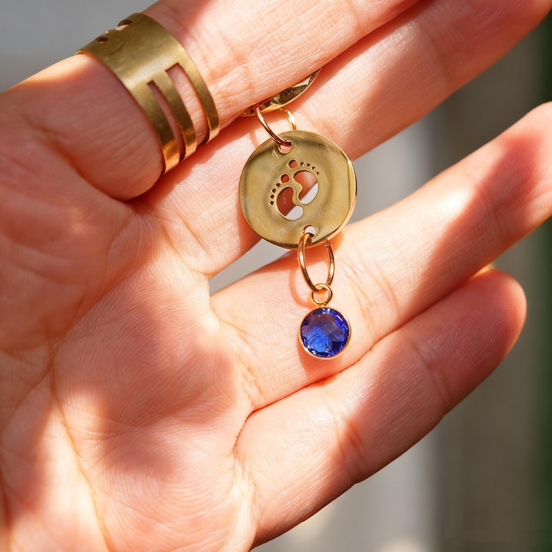 May include: A gold-colored pendant with a baby footprint design and a dangling blue gemstone. The pendant is attached to a gold ring. A gold ring with a cut-out design is on the finger.