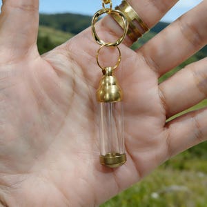 May include: A clear glass vial pendant with gold-colored metal accents. The vial is cylindrical and suspended from a gold-colored chain with a decorative loop. The pendant is held in a hand, with a blurred natural background.