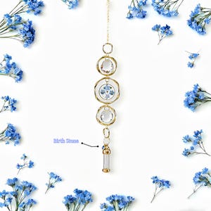 May include: A gold-colored, hanging birthstone ornament with three clear crystal orbs and a small glass vial. The ornament is suspended from a delicate chain and surrounded by blue flowers on a white background.