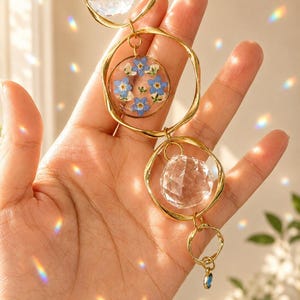 Puede incluir: Un adorno colgante con tres anillos de metal dorado conectados en una cadena vertical. Dos esferas de cristal transparente facetado cuelgan dentro de los anillos, y un dije circular de resina contiene flores nomeolvides azules prensadas.