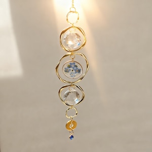 May include: A decorative hanging ornament with a gold chain and circular accents. The ornament features clear crystal spheres and a central section with blue flowers. A small blue gem hangs at the bottom, adding a touch of color.