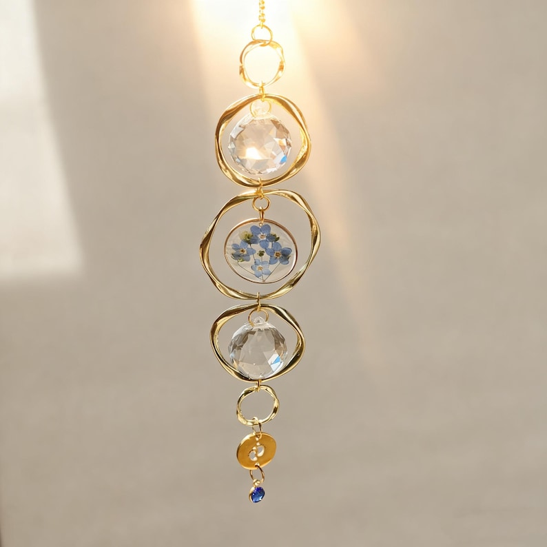 May include: A decorative hanging ornament with a gold chain and circular accents. The ornament features clear crystal spheres and a central section with blue flowers. A small blue gem hangs at the bottom, adding a touch of color.