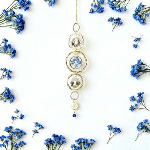 May include: A gold-colored hanging ornament with clear crystal-like spheres and blue flower accents. The ornament is suspended from a chain and has a small blue bead at the bottom. Blue flowers are scattered around the ornament on a white background.