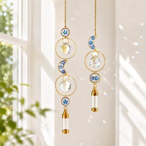 Könnte beinhalten: Zwei hängende Sonnenfänger mit goldenen Ketten, Reifen und Akzenten. Jeder verfügt über einen klaren Kristallkugel, einen Halbmond mit blauen floralen Details und eine kleine Glasampulle. Die Sonnenfänger hängen vor einem Fenster.