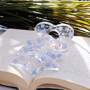 May include: Clear acrylic thumb book page holders with blue flower and gold leaf inclusions. The book accessories are resting on an open book with text. The holders are designed to keep pages open.