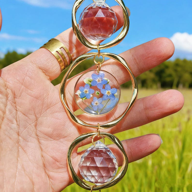 May include: A three-tiered gold-toned suncatcher with clear and red crystal spheres and a circular pendant with blue flowers. The suncatcher is held up against a backdrop of a blue sky and green field.
