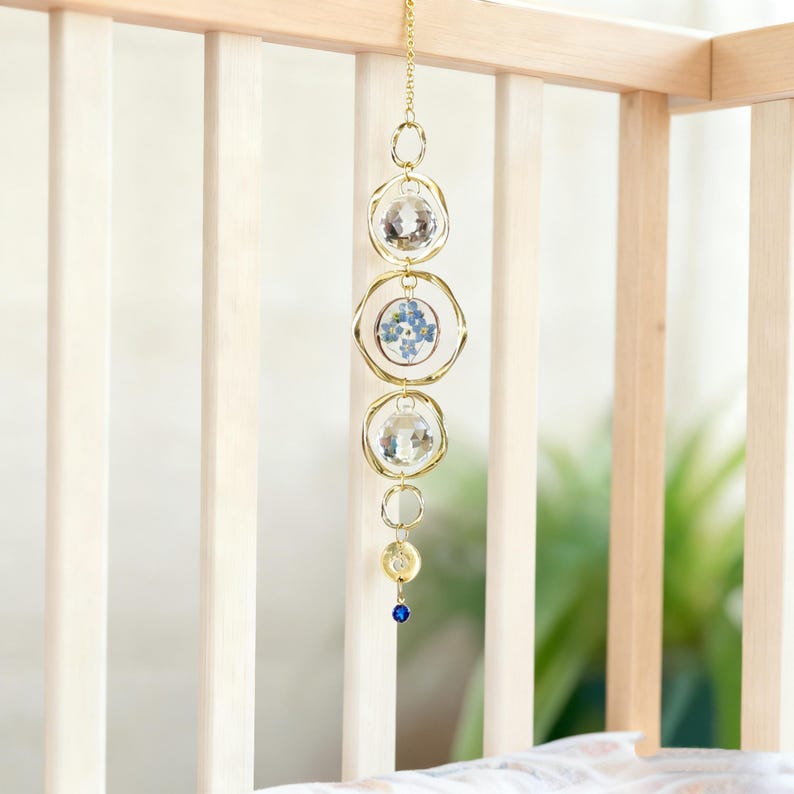 May include: A hanging mobile with a gold chain and circular frames. The mobile features clear crystal orbs and a circular frame with blue flowers. A small blue bead hangs at the bottom. The mobile is hanging in front of a wooden crib.