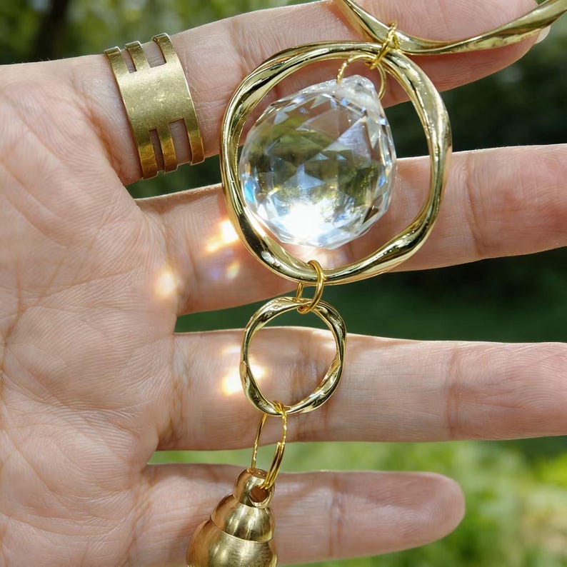 May include: A gold-toned necklace with a large, clear crystal pendant suspended within a circular frame. The necklace features a smaller, twisted gold ring and a bell-shaped charm. A gold ring with a geometric design is also visible.