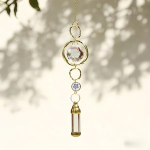 Könnte beinhalten: Ein hängender Sonnenfänger mit einem klaren Kristallkugel, goldfarbenen Metallringen und einem kleinen blauen Blumenakzent. Der Sonnenfänger hat auch ein klares Glasfläschchen am Boden, das an einer goldenen Kette hängt.
