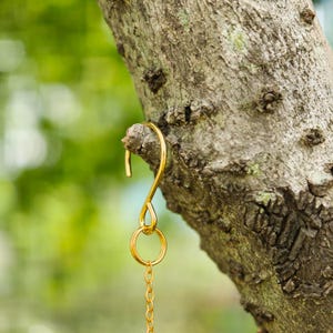 May include: A gold-colored metal hook hanging from a tree branch. The hook has a curved top, a circular ring, and a chain. The tree bark is textured and brown, with a blurred green background.