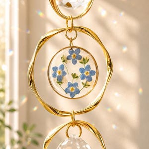 Puede incluir: Un atrapasoles colgante con dos esferas de cristal transparente facetado y un colgante circular central de resina. El colgante contiene flores de nomeolvides azules prensadas y follaje verde dentro de anillos de metal dorado con textura retorcida.