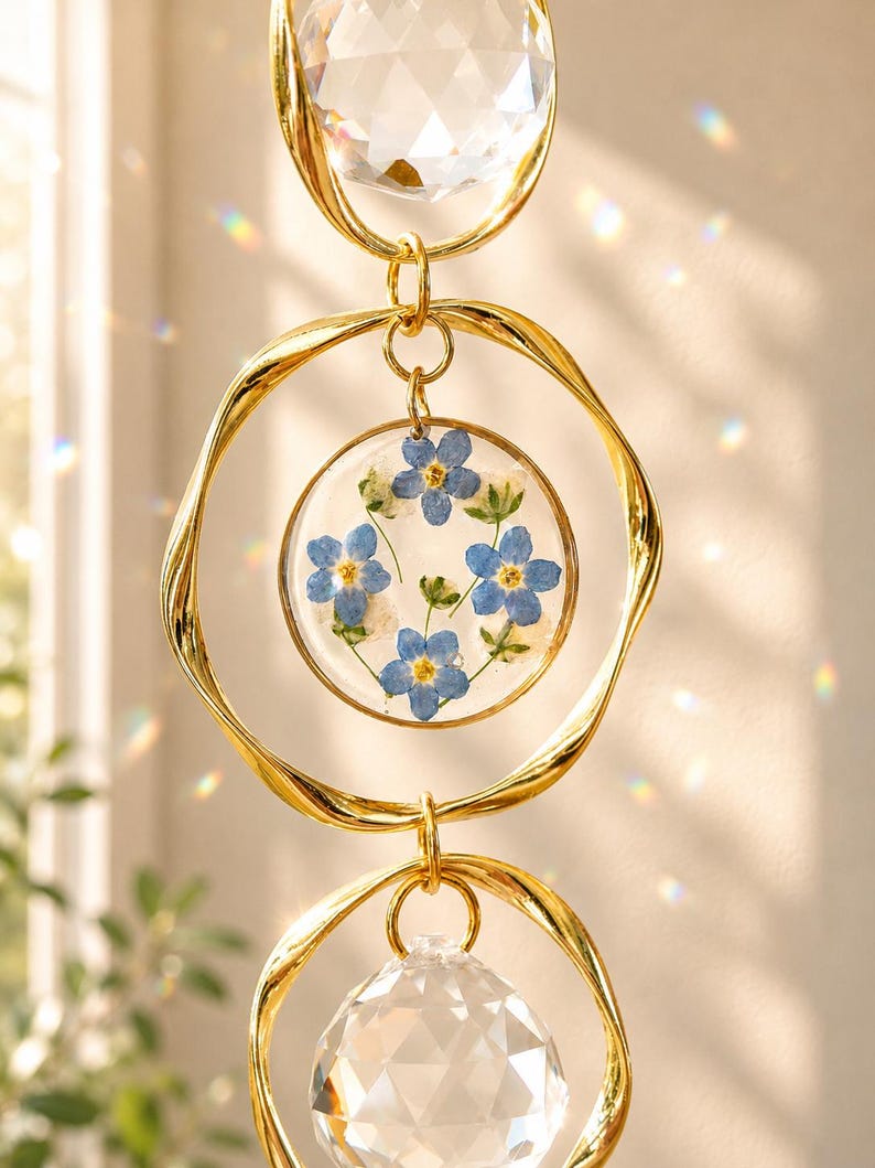 Puede incluir: Un atrapasoles colgante con dos esferas de cristal transparente facetado y un colgante circular central de resina. El colgante contiene flores de nomeolvides azules prensadas y follaje verde dentro de anillos de metal dorado con textura retorcida.