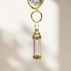 May include: A gold-toned pendant with a clear, cylindrical vial suspended below a faceted crystal. The vial is encased in gold metal at the top and bottom, with a clear glass center. The pendant is designed with a heart-shaped crystal.