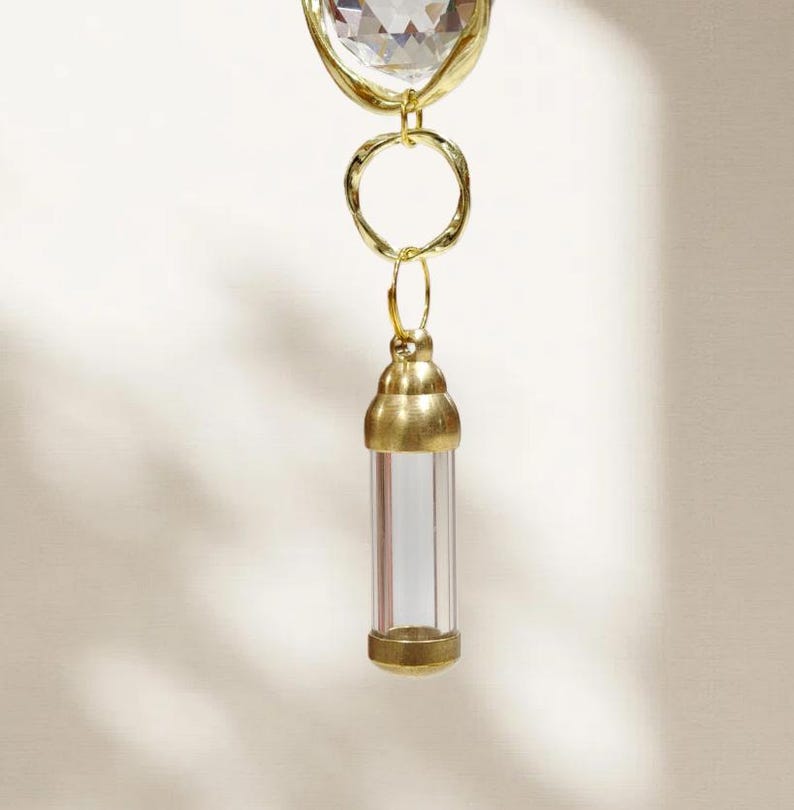 May include: A gold-toned pendant with a clear, cylindrical vial suspended below a faceted crystal. The vial is encased in gold metal at the top and bottom, with a clear glass center. The pendant is designed with a heart-shaped crystal.