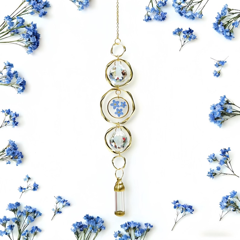 May include: A gold-colored suncatcher with three crystal orbs and a small glass vial at the bottom. The center orb contains blue flowers. The suncatcher is surrounded by blue flowers on a white background.