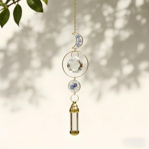Könnte beinhalten: Ein himmlischer Sonnenfänger mit goldener Kette, Halbmond und kreisförmigen Elementen. Das Design beinhaltet einen klaren Kristallkugel und kleine blaue Blumen. Am unteren Ende hängt ein klares, zylindrisches Glasfläschchen.