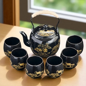 May include: A black Japanese tea set with a teapot and six matching cups. The teapot has a woven handle and gold-colored landscape designs. The cups and teapot have a glossy finish.