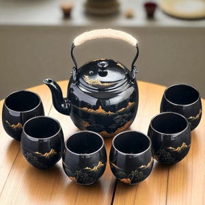 Puede incluir: Un juego de té de cerámica negra con una tetera y seis tazas a juego. La tetera tiene un asa trenzada y un pico. El juego está decorado con un diseño de paisaje dorado. Las tazas son redondas y también presentan el paisaje dorado.