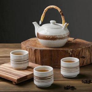 Könnte beinhalten: Ein Keramik-Teeservice mit einer weißen Teekanne mit Bambusgriff und drei passenden Tassen. Die Teekanne hat ein braunes Streifendesign und die Tassen braune horizontale Streifen. Das Teeservice steht auf einer Holzoberfläche.