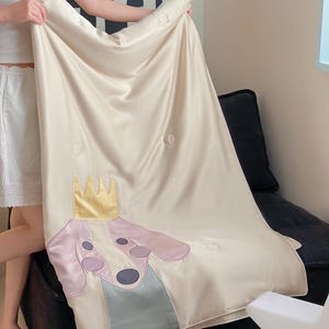 May include: Cream-colored satin blanket featuring a cartoon dog wearing a gold crown. The dog has a pink head, purple ears, and a blue-gray collar. The blanket has a quilted design with circular stitching.