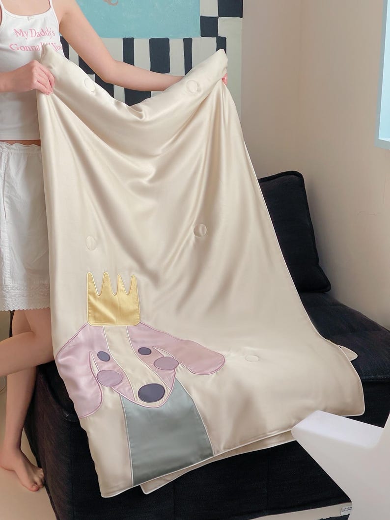 May include: Cream-colored satin blanket featuring a cartoon dog wearing a gold crown. The dog has a pink head, purple ears, and a blue-gray collar. The blanket has a quilted design with circular stitching.