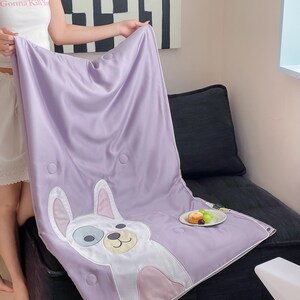 May include: A lavender satin blanket featuring a cartoon dog design with white, blue, and pink accents. The blanket has a quilted pattern with circular stitching. A small plate of fruit sits nearby.