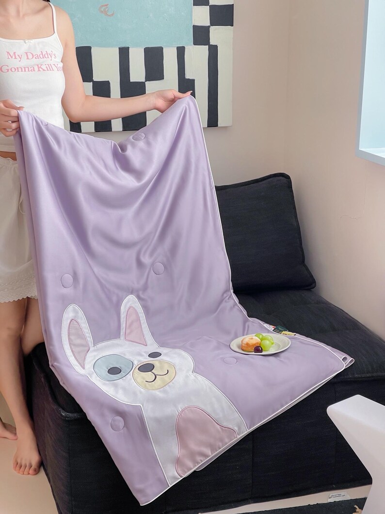May include: A lavender satin blanket featuring a cartoon dog design with white, blue, and pink accents. The blanket has a quilted pattern with circular stitching. A small plate of fruit sits nearby.