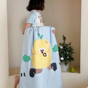 May include: A light blue blanket featuring a cartoon lemon character with green mittens and black shoes. The lemon has a smiling face and is running. The blanket is held up by a person.
