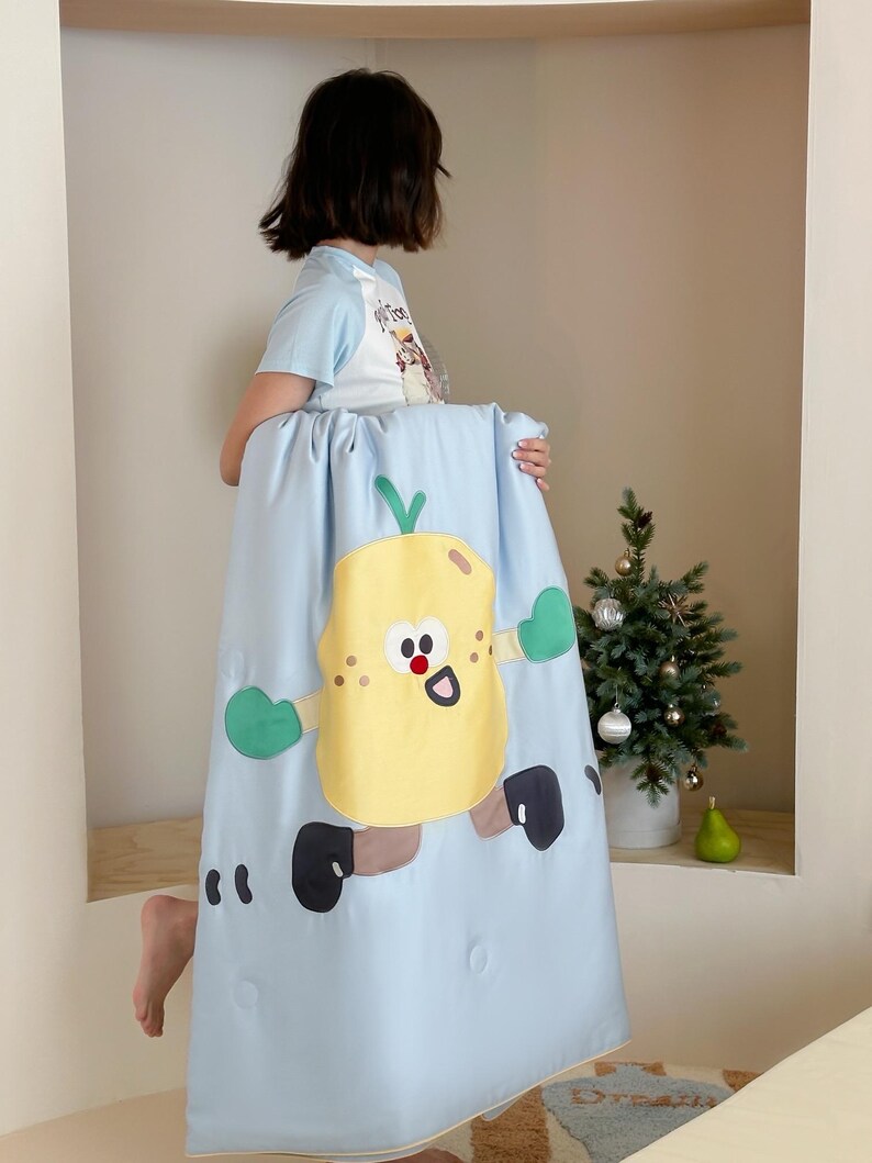 May include: A light blue blanket featuring a cartoon lemon character with green mittens and black shoes. The lemon has a smiling face and is running. The blanket is held up by a person.