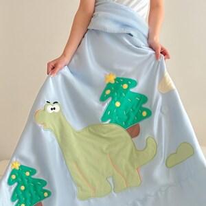 May include: Light blue satin blanket featuring a green dinosaur and two green Christmas trees with yellow stars. The blanket has a cloud design and is being held up by a person wearing a white t-shirt with the text "My Daddy's Gonna Kill You".
