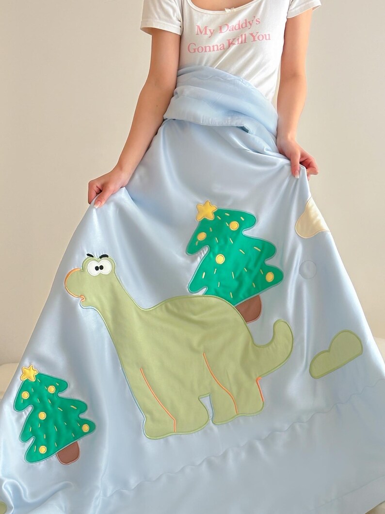 May include: Light blue satin blanket featuring a green dinosaur and two green Christmas trees with yellow stars. The blanket has a cloud design and is being held up by a person wearing a white t-shirt with the text "My Daddy's Gonna Kill You".