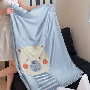 May include: A light blue blanket with a cartoon bear face and striped shirt design. The bear has a cream-colored face, orange cheeks, and a blue nose. The blanket has a smooth, satin-like texture and is decorated with round, light blue buttons.