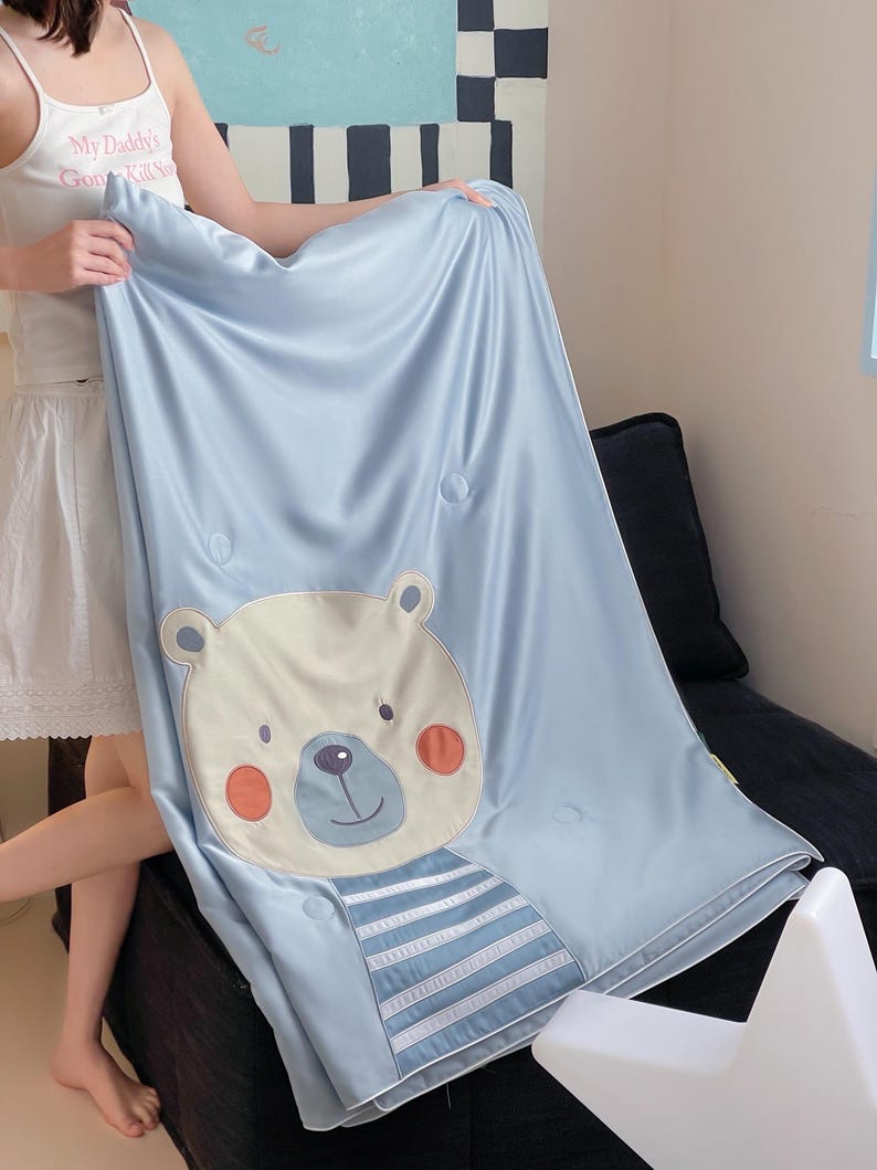 May include: A light blue blanket with a cartoon bear face and striped shirt design. The bear has a cream-colored face, orange cheeks, and a blue nose. The blanket has a smooth, satin-like texture and is decorated with round, light blue buttons.